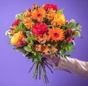 Blombukett med bland nnat rosor, gerbera, nejlikor och alstroemeria tillsammans med grönt. Blomfärger rött, orange, gult.