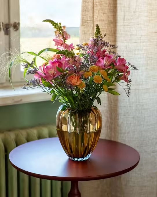Beställ ditt blomsterbud hos Interflora! Interfloras färgsprakande augustibukett, med lejongap, alstroemeria, nejlika, krysantemum och gröna blad. Rosa, gult, orange, grönt. Här en bild på buketten i en fin vas som står på ett runt bord vid ett fönster. En gardin bakom bordet syns vid ena sidan av fönstret.