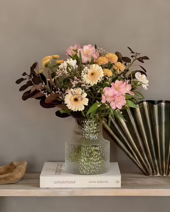 Oktoberbuketten från Interflora. Här, en bild på buketten i en glasvas som står på en beige bok som ligger på en beige hylla. Grön dragspelsformad dekoration till höger, trädekoration till vänster om vasen. I buketten: germini, krysantemum, alstroemeria, limonium, perukbuske, dekorationsgrönt.