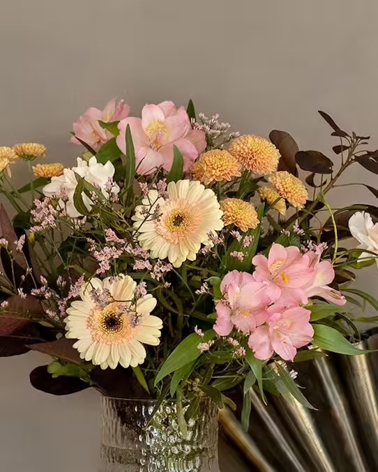 Bild på blommorna i Interfloras oktoberbukett, mot en beigebrun bakgrund. Blommorna står i en glasvas. Blommor: germini, krysantemum, alstroemeria, limonium, perukbuske, dekorationsgrönt